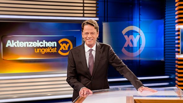 Moderator Rudi Cerne steht im Aktenzeichen XY-Studio und lacht in die Kamera. - Foto: ZDF / Nadine Rupp