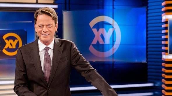 Moderator Rudi Cerne steht im Aktenzeichen XY-Studio und lacht in die Kamera. - Foto: ZDF / Nadine Rupp