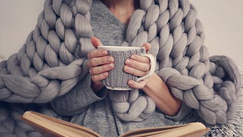 Frau trinkt Tee, liest und ist mit einer Decke zugedeckt. - Foto: iStock/Albina Gavrilovic