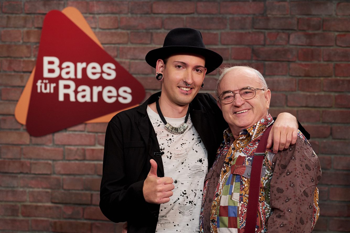 'Bares für Rares': Die Händler Fabian Kahl und Ludwig Hofmaier stehen vor dem Sendungslogo und lächeln in die Kamera.