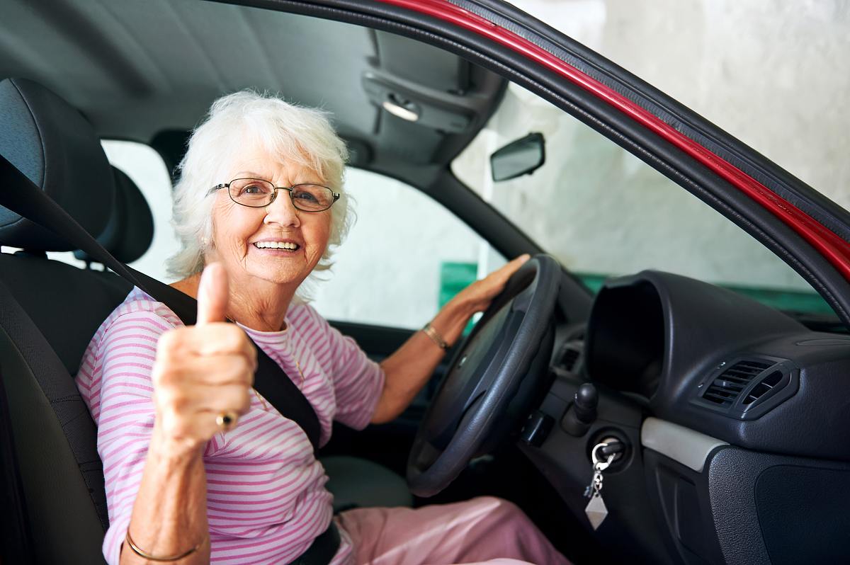 Eine lächelnde ältere Dame sitzt am Steuer ihres Wagens und macht ihrem Daumen hoch.