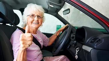 Eine lächelnde ältere Dame sitzt am Steuer ihres Wagens und macht ihrem Daumen hoch. - Foto: iStock / warrengoldswain