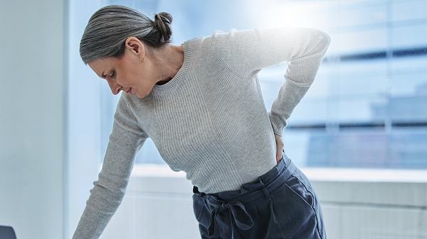 Mittelalter Frau mit hellem Pullover fasst sich schmerzerfüllt an den Rücken - Foto: iStock/PeopleImages