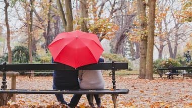 Paar sitzt unter einem Regenschirm auf einer Bank. 