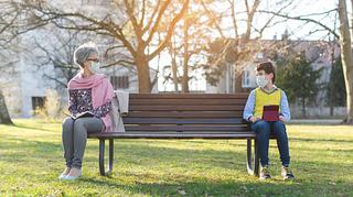 Eine Frau und ein Junge sitzen mit Abstand auf einer Parkbank. - Foto: iStock/ kzenon