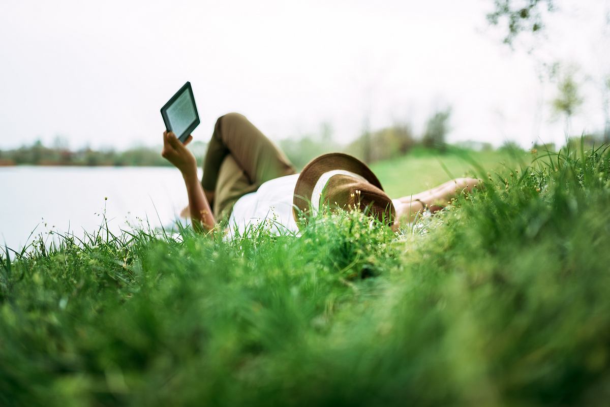 Frau liegt an einem See in der Wiese und liest auf einem E-Reader