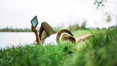 Frau liegt an einem See in der Wiese und liest auf einem E-Reader - Foto: nortonrsx / iStock