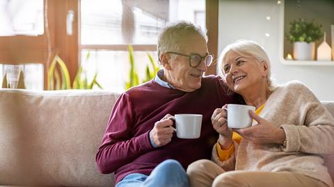 Ein älteres Ehepaar hat es sich mit Kaffeetassen auf der Couch gemütlich gemacht. - Foto: iStock / Piksel