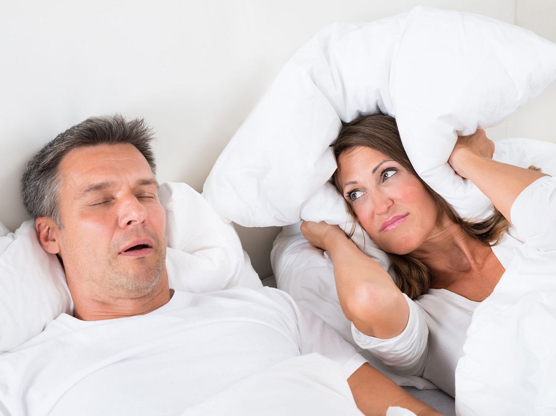 Paar liegt im Bett, Mann schnarcht und hat Atemaussetzer, Frau hält sich die Ohren zu.