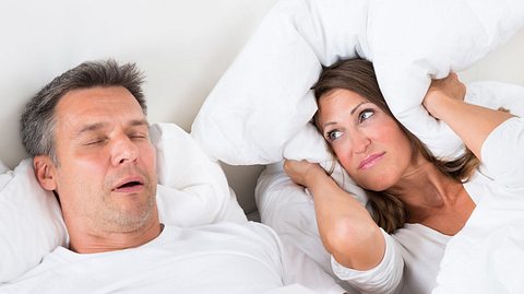 Paar liegt im Bett, Mann schnarcht und hat Atemaussetzer, Frau hält sich die Ohren zu. - Foto: AndreyPopov / iStock
