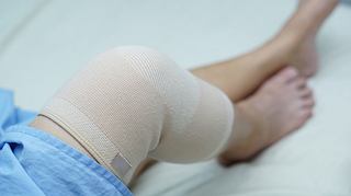 Frau liegt mit einem bandagierten Knie auf einem Bett. - Foto: sasirin pamai / iStock