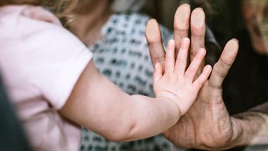 Kleines Mädchen drückt ihre Hand gegen eine Scheibe an Omas Hand. - Foto: RyanJLane / iStock